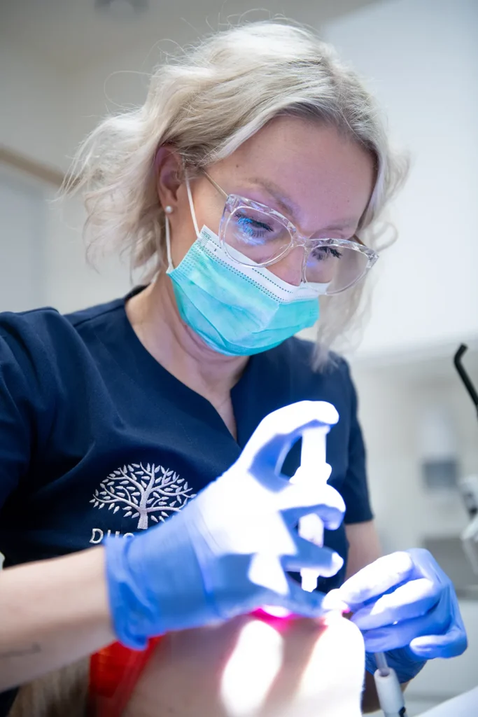 Specjalistka kliniki DUNIQUE w granatowym uniformie z logo firmy, w masce ochronnej i okularach, skoncentrowana podczas precyzyjnego zabiegu stomatologicznego z wykorzystaniem nowoczesnego sprzętu, symbolizując profesjonalizm i dbałość o najwyższą jakość leczenia.